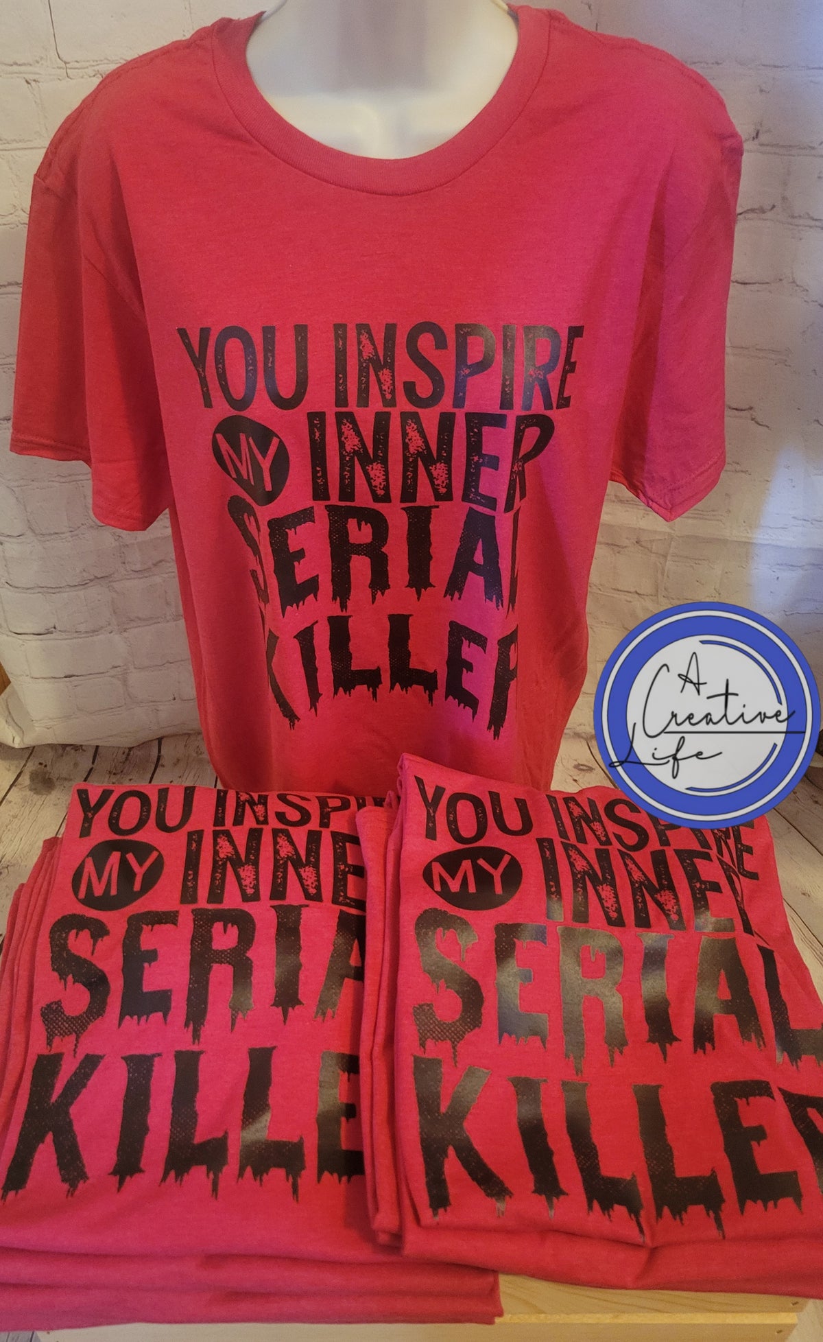 Two red t-shirts displayed on a mannequin, one with the text 'YOU INSPIRE ME' and the other with the text 'INNER SERIAL KILLER'. Both shirts have a black graphic logo on the front.
