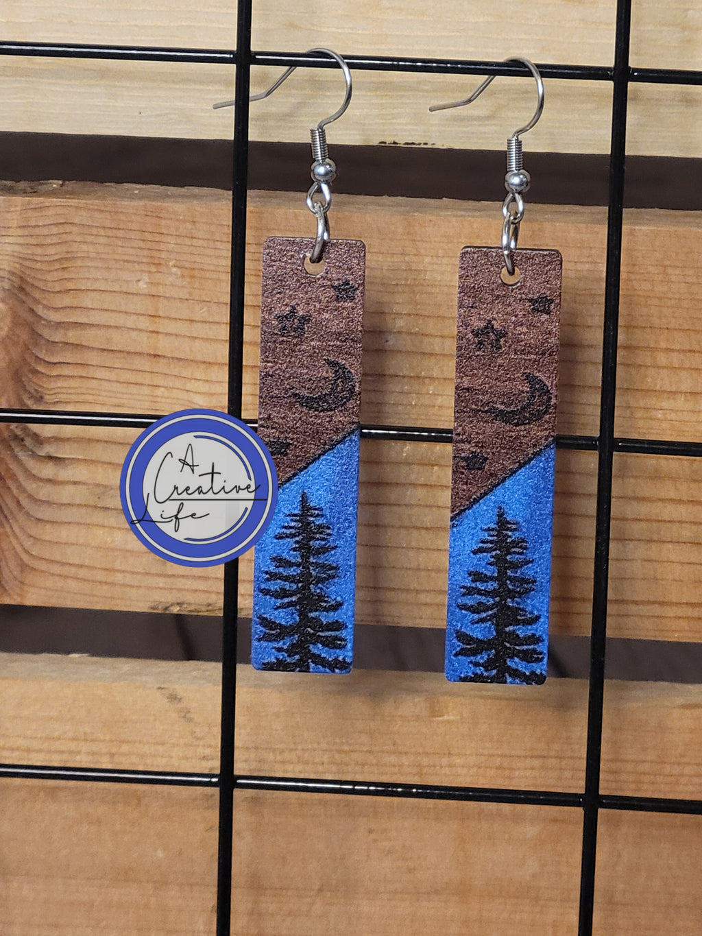 A pair of earrings with a night sky pattern, featuring trees and stars, displayed on a wooden plank.