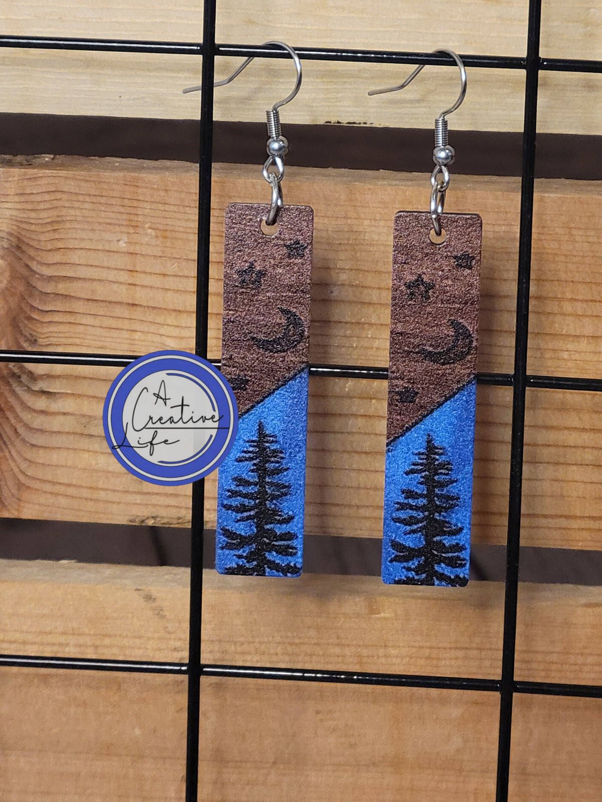 A pair of earrings with a night sky pattern, featuring trees and stars, displayed on a wooden plank.
