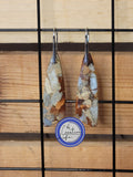 A pair of boho style earrings with an earth tone appearance, hanging from stainless steel hooks with an 18K white gold finish, displayed against a wooden backdrop.