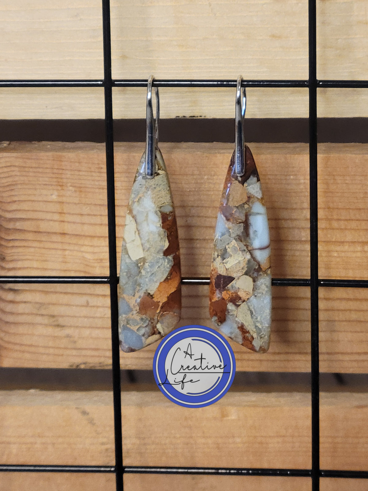 A pair of boho style earrings with an earth tone appearance, hanging from stainless steel hooks with an 18K white gold finish, displayed against a wooden backdrop.