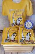 A yellow t-shirt with a graphic print of gnomes and the text 'Grateful, Thankful, Blessed' displayed on a wooden surface.