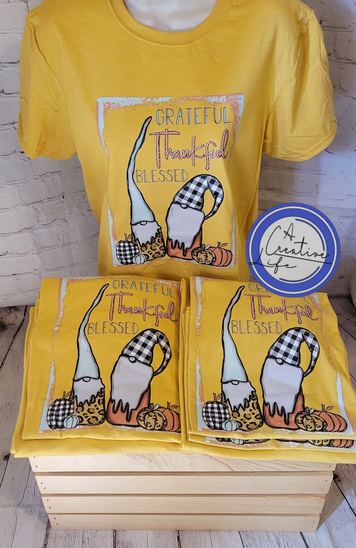 A yellow t-shirt with a graphic print of gnomes and the text 'Grateful, Thankful, Blessed' displayed on a wooden surface.