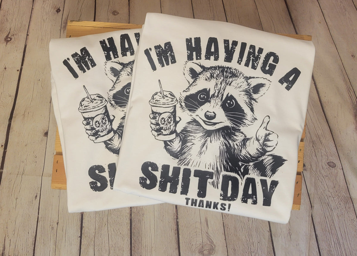 Two white t-shirts with a cartoon raccoon and text, one displayed in front view and the other in side view, both on a wooden surface.
