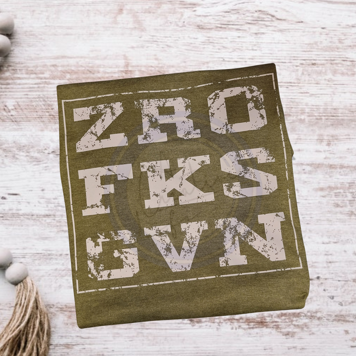 A military heather green T-shirt with text 'ZRO FKS GVN' printed in a distressed white font, displayed on a wooden surface.
