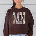 Person wearing a brown sweatshirt with 'MN' printed on it with a camo wooden background