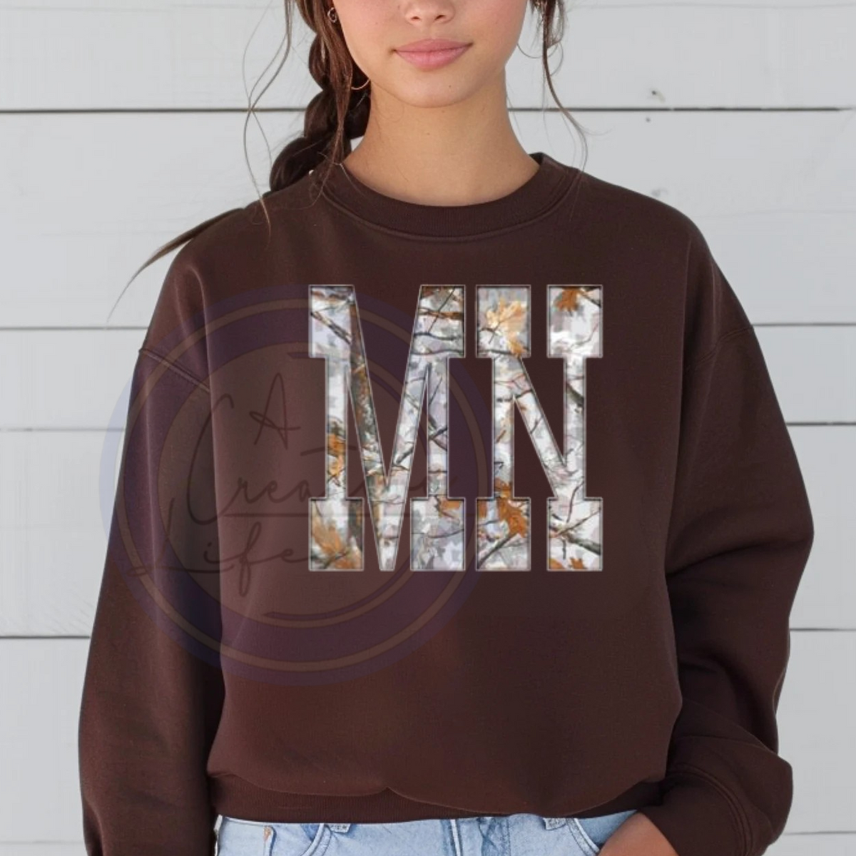 Person wearing a brown sweatshirt with 'MN' printed on it with a camo wooden background