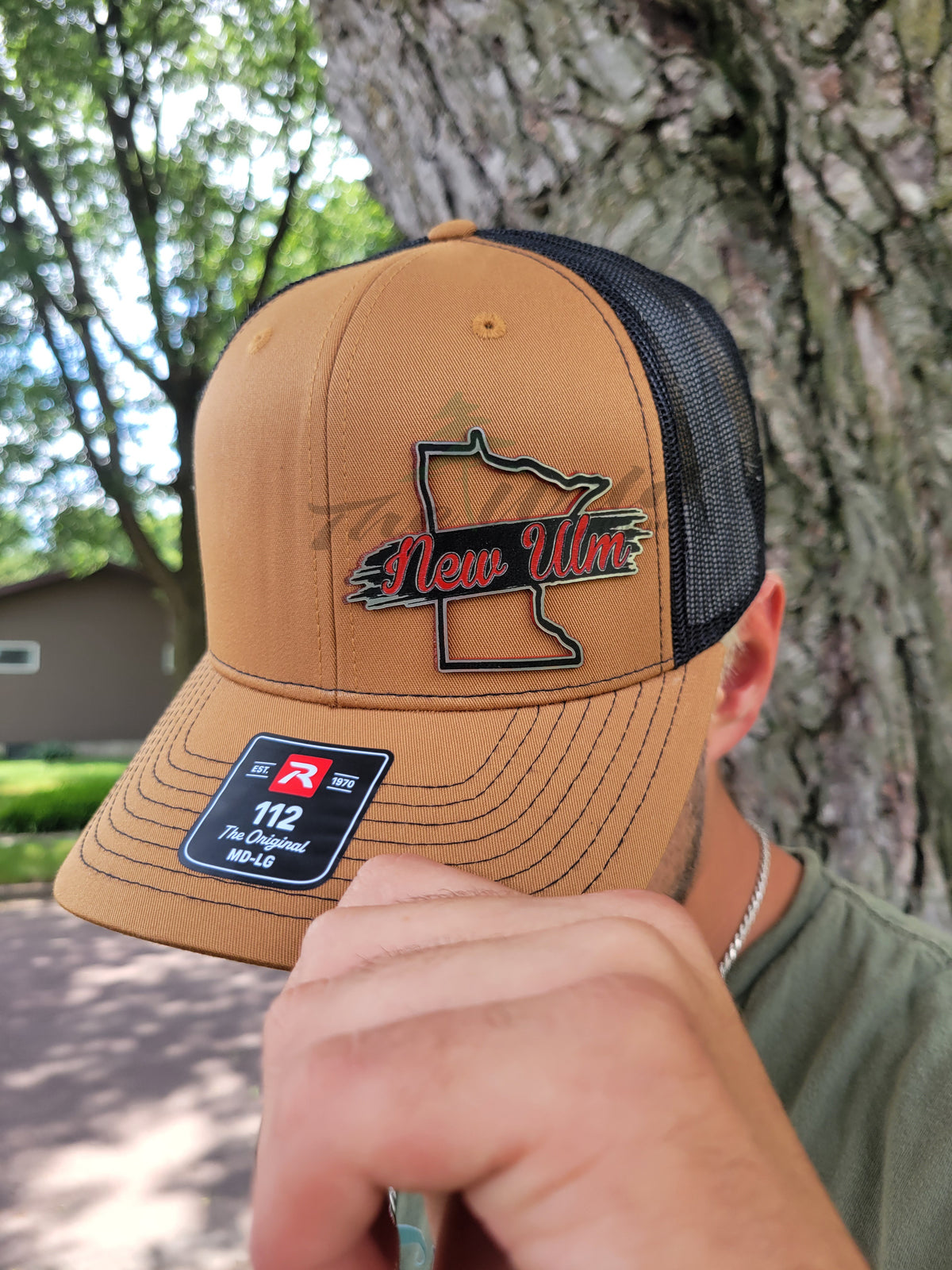 Brown cap with black mesh back and 'New Ulm' logo worn by a person, with a tree and building in the background.