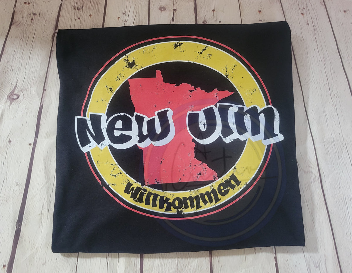Black t-shirt with a red and yellow New Ulm Willkommen graphic design on a wooden surface