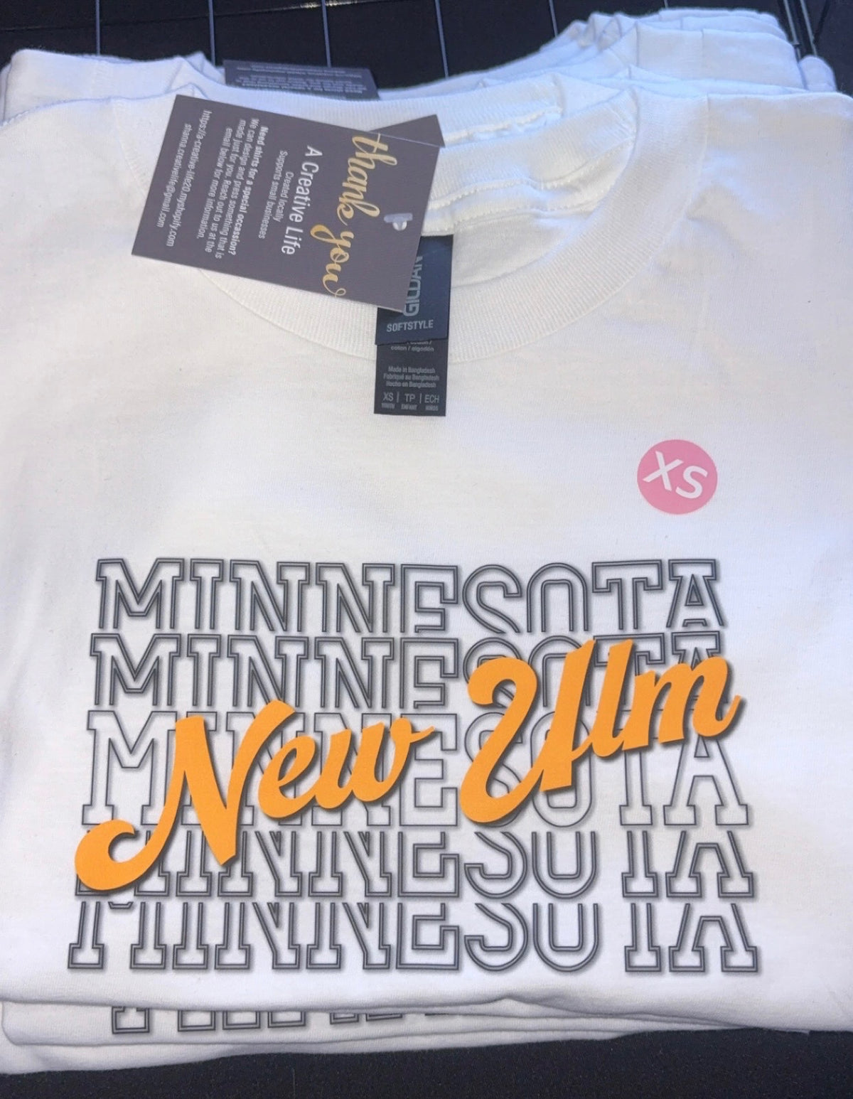 A folded white crewneck sweatshirt featuring a stacked 'MINNESOTA' graphic in gray outlined letters, with 'New Ulm' in orange script across the front and a small pink circular 'xs' size sticker on the upper right chest.