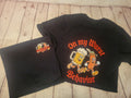 A black graphic t-shirt laid flat on a light wood plank surface, with a matching black folded garment placed underneath. The shirt features a large cartoon-style beer mug and sausage character graphic with the text 'On my Wurst Behavior' in orange.