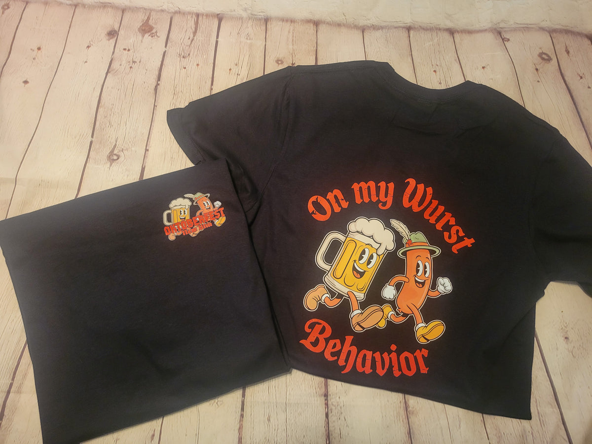 A black graphic t-shirt laid flat on a light wood plank surface, with a matching black folded garment placed underneath. The shirt features a large cartoon-style beer mug and sausage character graphic with the text 'On my Wurst Behavior' in orange.