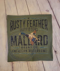 Green folded tshirt with 'Rusty Feather Outfitters Mallard Brand Co. American Waterfowl' text and duck illustration on a wooden surface.