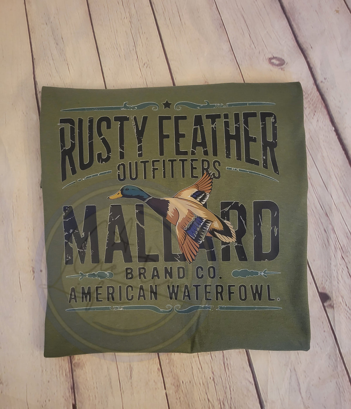 Green folded tshirt with 'Rusty Feather Outfitters Mallard Brand Co. American Waterfowl' text and duck illustration on a wooden surface.