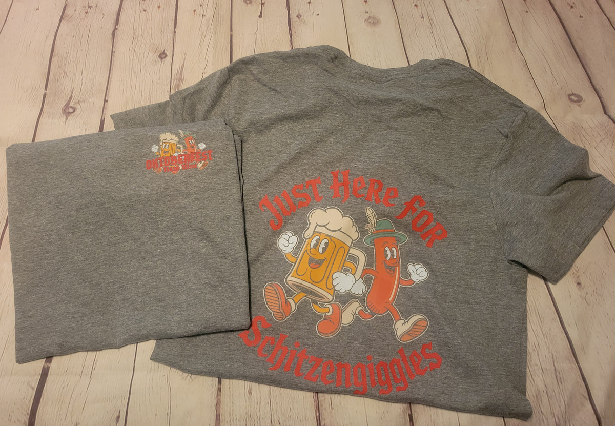 A heather gray t-shirt laid flat on a light wood plank background, showing both the front and back views. The back features a colorful graphic of a beer mug and a hot dog character with the text 'Just Here For Schitzengiggles'.