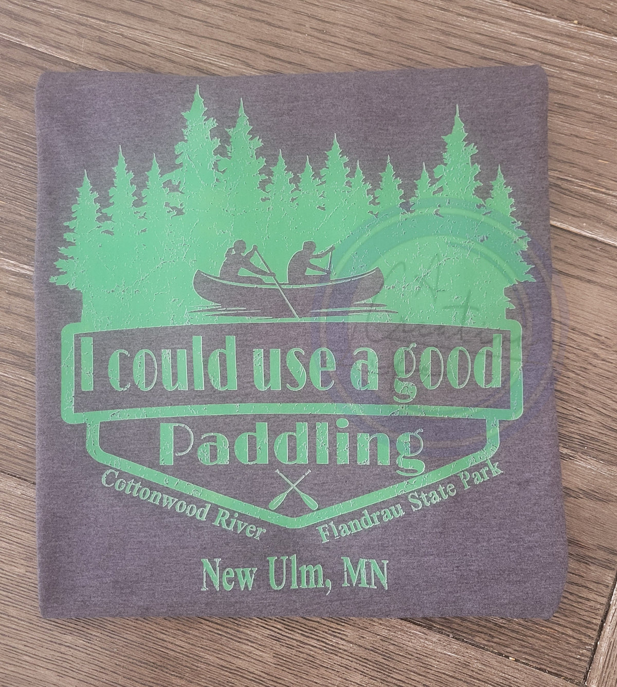 Tshirt with green text about I could use a good paddling in reference to the cottonwood in New Ulm MN  and graphics on a wooden surface