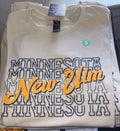 A folded cream-colored t-shirt featuring a 'New Ulm Minnesota' graphic with layered 'MINNESOTA' text in dark gray outline and 'New Ulm' in orange script. A small green circular 'S' size label is visible on the chest.