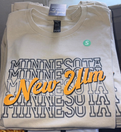 A folded cream-colored t-shirt featuring a 'New Ulm Minnesota' graphic with layered 'MINNESOTA' text in dark gray outline and 'New Ulm' in orange script. A small green circular 'S' size label is visible on the chest.