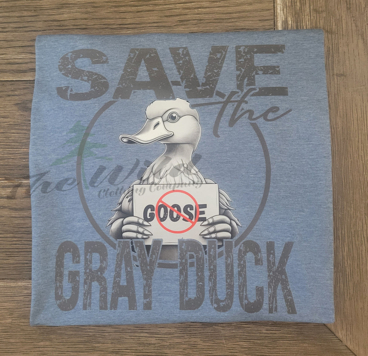 heather blue shirt with graphic of a duck holding a sign and text 'Save the Gray Duck' on a wooden surface.