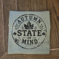 A folded light sage green tshirt, featuring a distressed black circular stamp design that reads 'AUTUMN STATE OF MIND' with maple leaves, placed on a dark wood background.