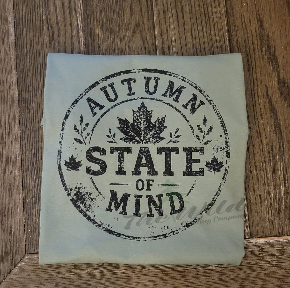 A folded light sage green tshirt, featuring a distressed black circular stamp design that reads 'AUTUMN STATE OF MIND' with maple leaves, placed on a dark wood background.