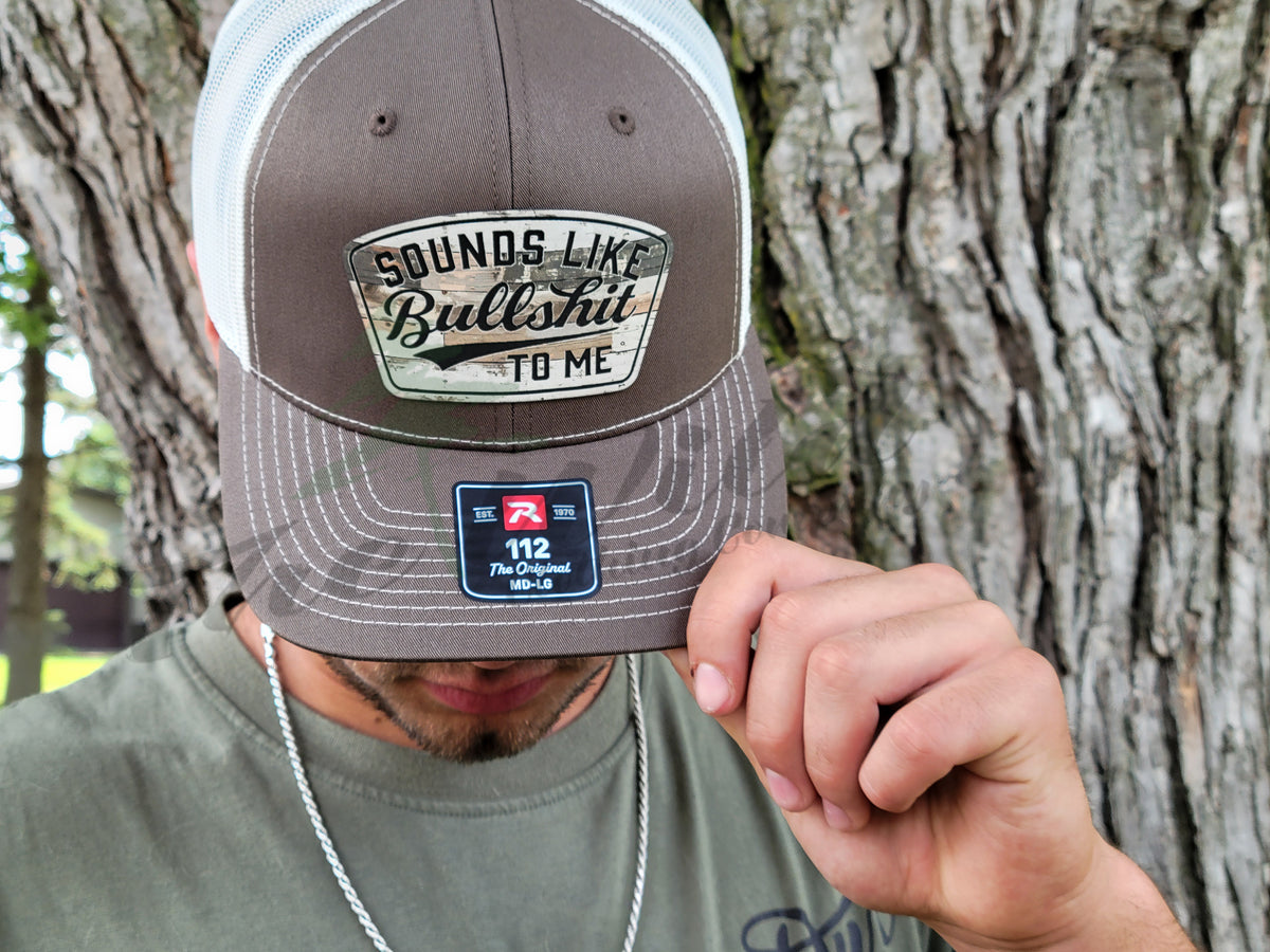 Person wearing a cap with text and branding, held up to the camera against a tree background
