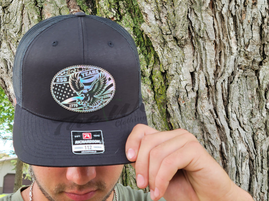 Person holding a black cap with a holographic 250 years American eagle decorative emblem against a tree background